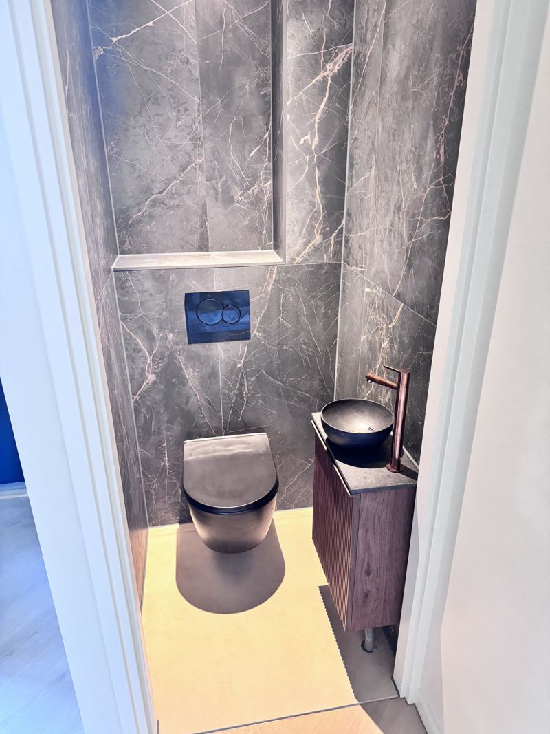 Een smalle toiletruimte met een mat zwart hangtoilet en een zwarte waskom op een notenhouten meubel. Een betegelde nis boven het reservoir geeft opbergruimte zonder kastje.