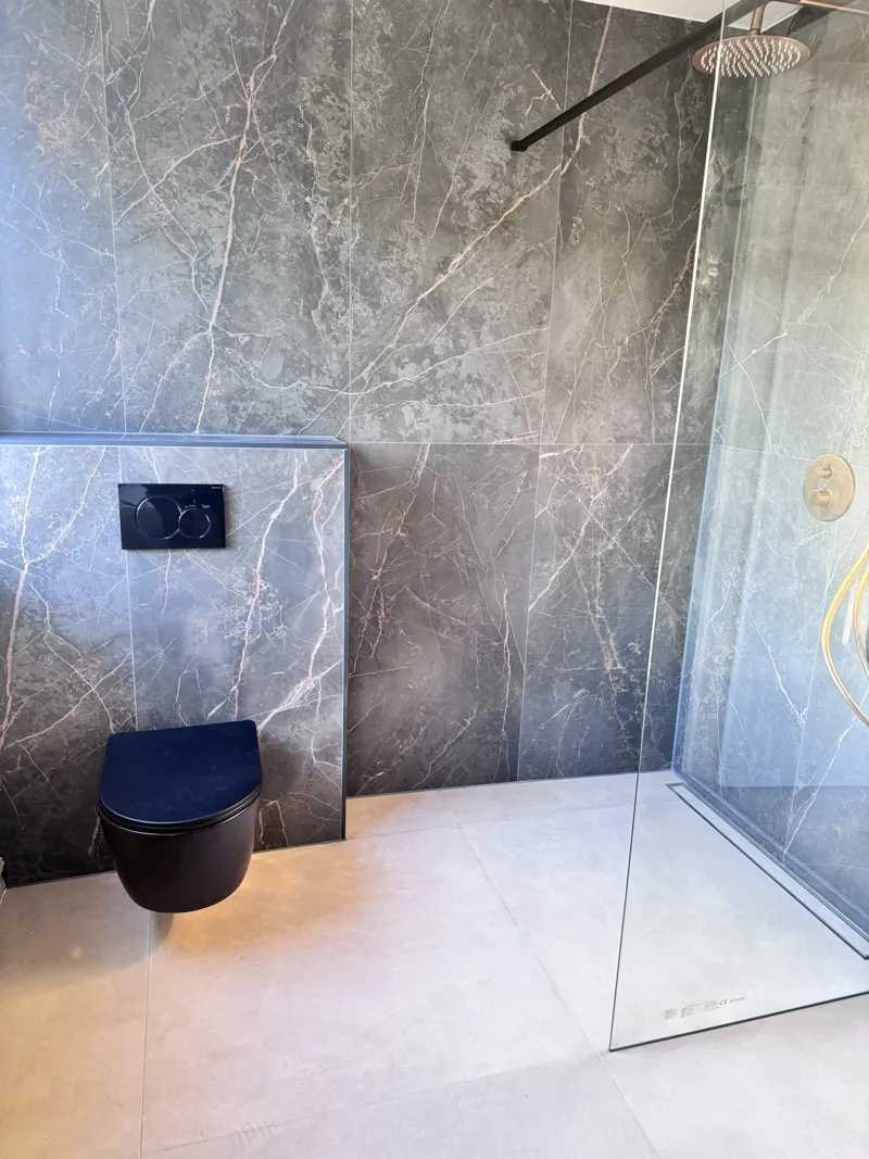 Een inloopdouche achter één glazen wand, met regendouche en thermostaat in geborsteld goud. Het toilet hangt voor een ingebouwd reservoir dat in dezelfde tegel is afgewerkt.